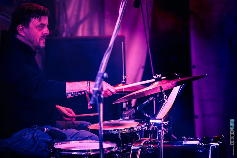 A male drummer playing a drum kit during a live music performance under purple stage lighting.