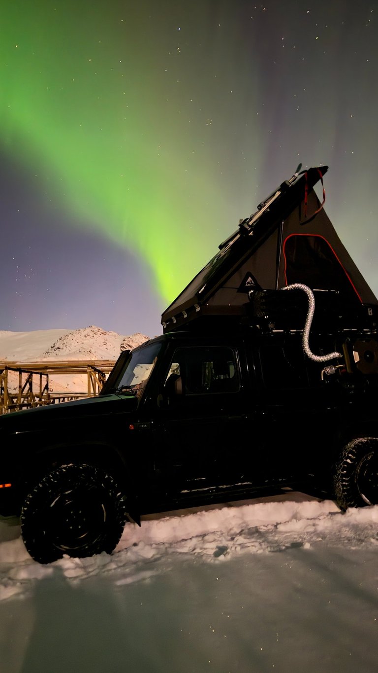 Black off-road truck with a roof top tent camping under the green Northern Lights in a snowy landscape.
