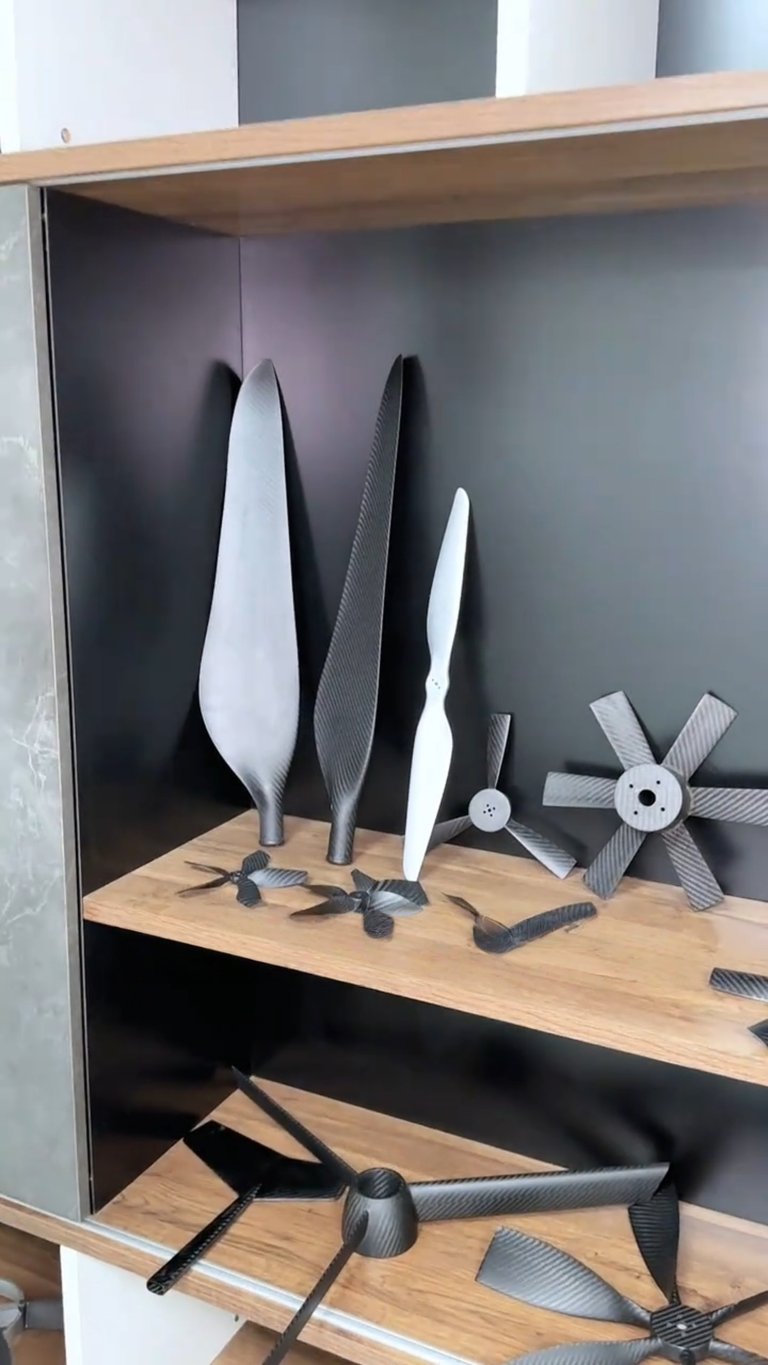 Collection of carbon fiber drone propellers and multi-blade rotors displayed on wooden shelves.