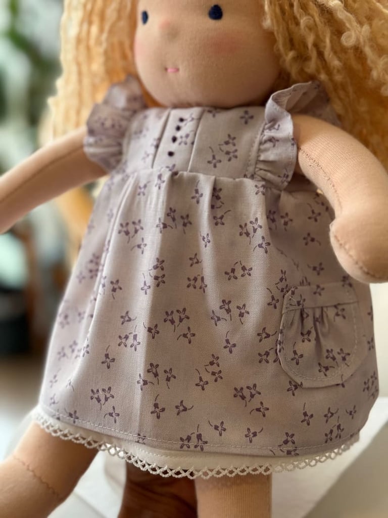 Muñeca Waldorf hecha a mano que lleva un vestido de algodón color lavanda con estampado floral