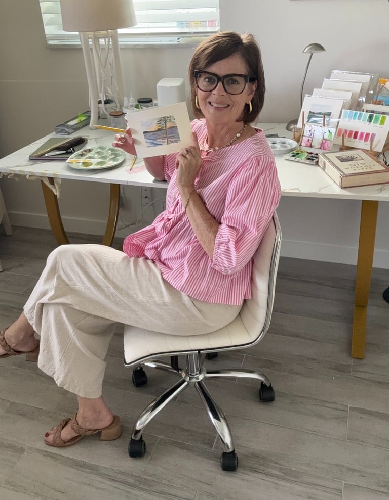 A smiling artist in a pink striped shirt holds her watercolor landscape painting at her bright home studio desk.