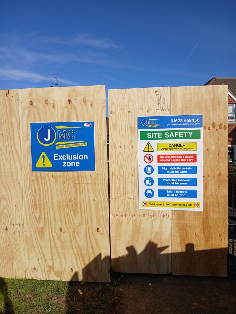 Site safety and exclusion zone signs posted on a plywood construction hoarding under a clear blue sky.