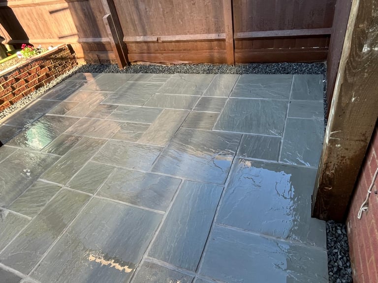 Wet grey sandstone patio tiles installed in a backyard with a wooden fence and brick wall.