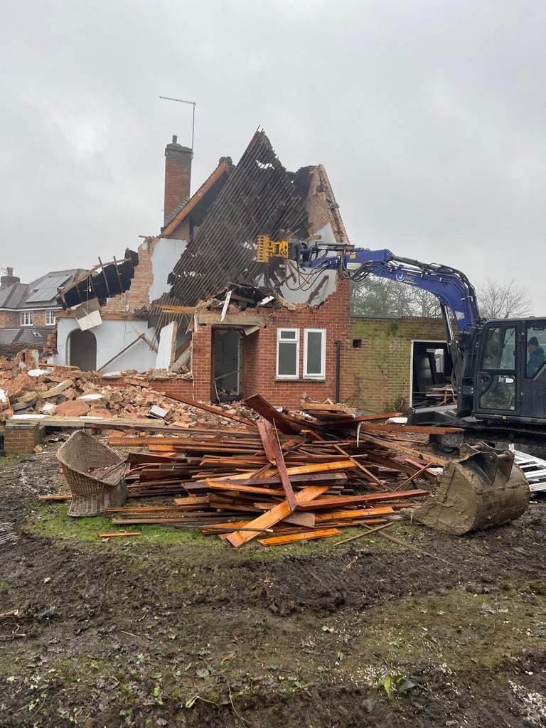 A blue hydraulic excavator demolition machine tearing down a damaged brick house with debris and wood piles.