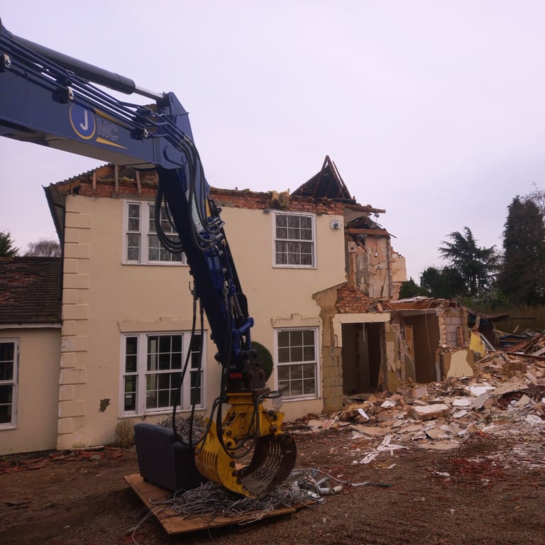 Blue excavator arm demolishing a two-story residential house with debris and rubble on the construction site.
