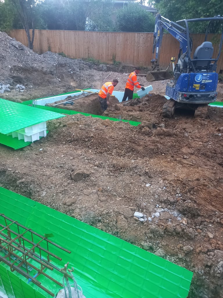 Construction workers installing green insulated concrete forms for building foundations with a mini excavator.