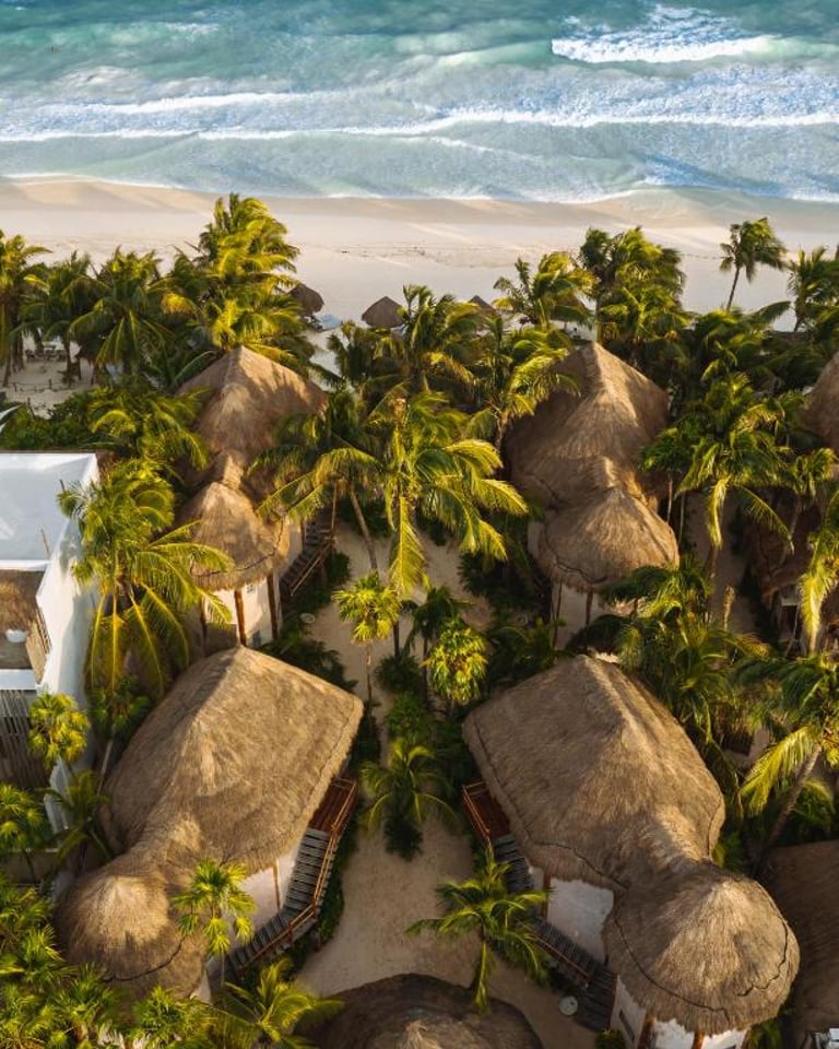 Aerial view of luxury thatched roof bungalows and palm trees on a white sand beach in Tulum, Mexico.