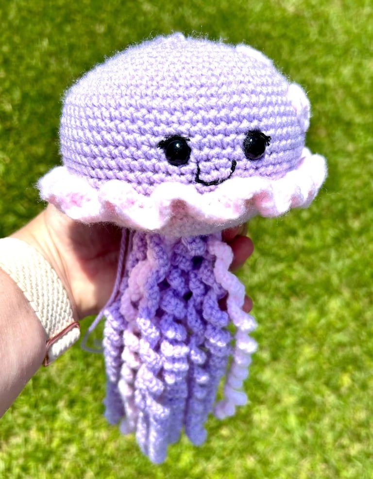 Handmade purple and pink crochet jellyfish amigurumi toy with curly tentacles held outdoors over green grass.