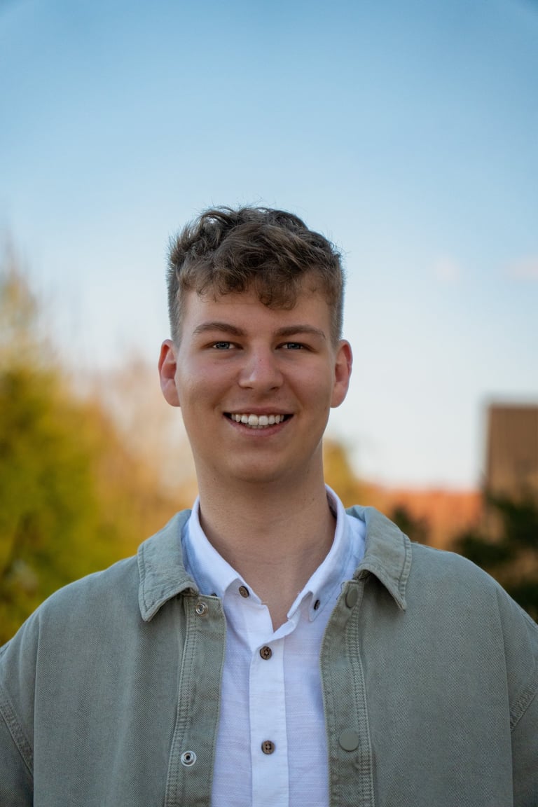 Ein lächelnder junger Mann mit blondem Haaren, der eine grüne Jacke über einem weißen Hemd trägt.