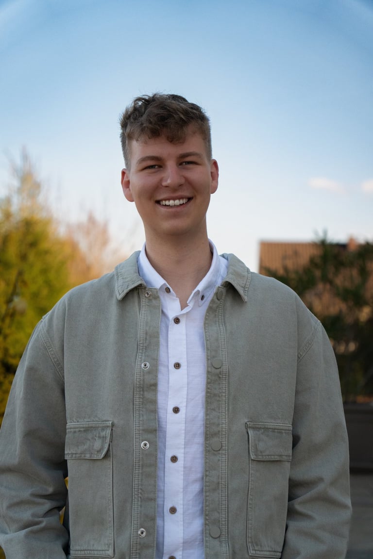 Ein lächelnder junger Mann mit blondem Haaren, der eine grüne Jacke über einem weißen Hemd trägt.