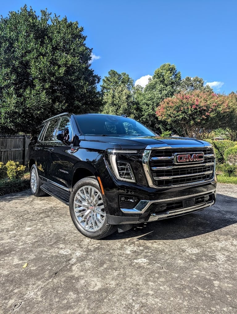 A black GMC Yukon Denali SUV parked on a concrete driveway with a lush green tree background.