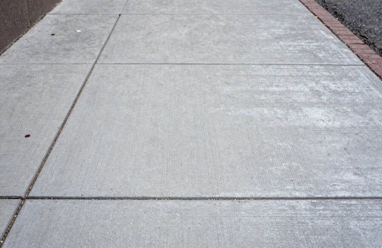Close-up texture photograph of a perfectly executed broom-finish concrete sidewalk in a North American / US urban setting, showing the sturdy silver surface.