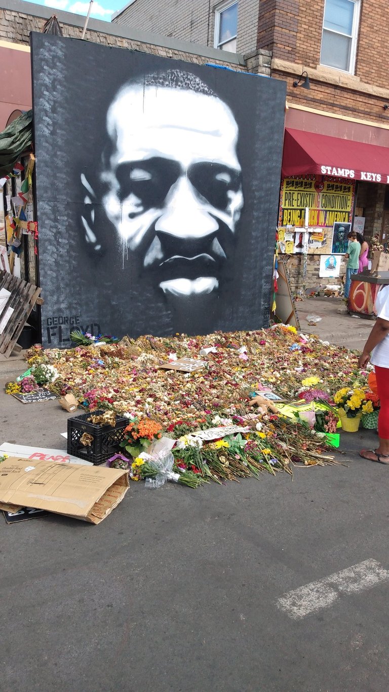 A black and white mural of George Floyd sits behind a large pile of memorial flowers.