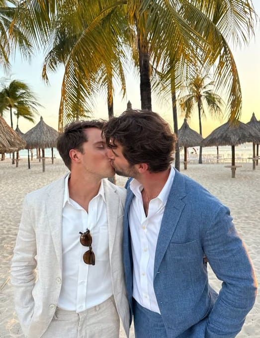 A gay couple in linen suits sharing a romantic kiss on a tropical beach at sunset with palm trees.