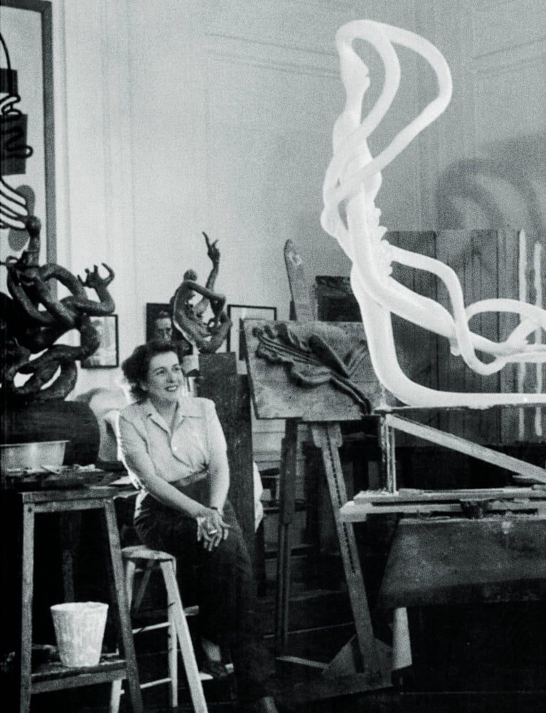 Artist Maria Martins smiles in her studio surrounded by large abstract modernist sculptures.