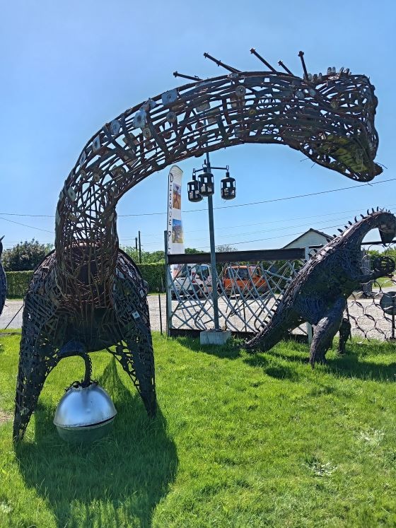 A large outdoor metal scrap sculpture of a long-necked dinosaur standing on a sunny green lawn.
