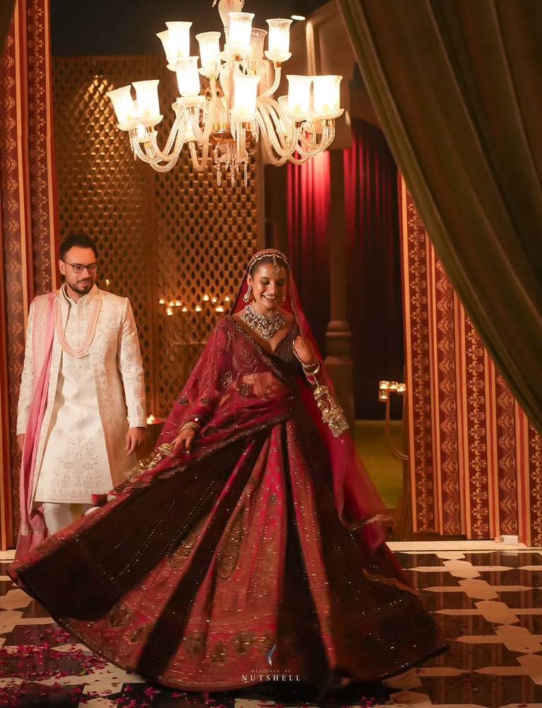 Indian bride twirling in a maroon bridal lehenga while the groom watches under a crystal chandelier.
