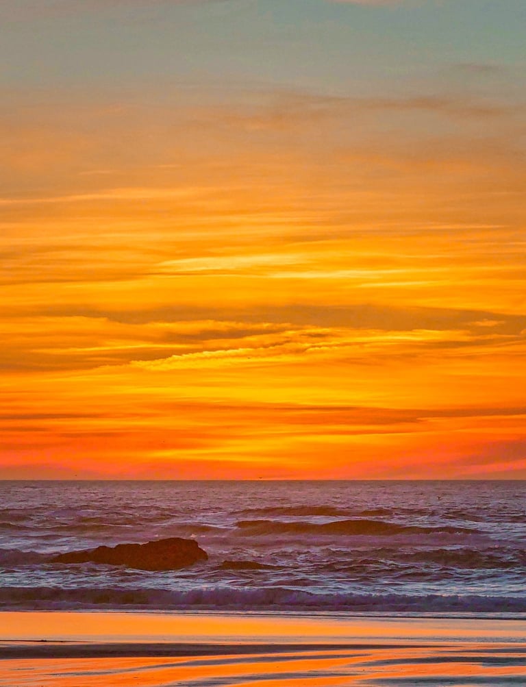 Vibrant orange sunset sky over ocean waves with a rocky beach shoreline at dusk.