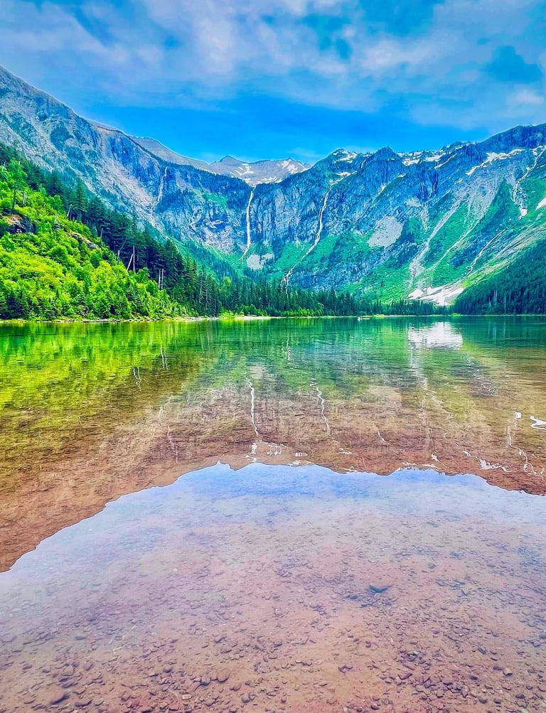 Clear alpine lake reflecting rugged mountains and green forests under a blue sky.