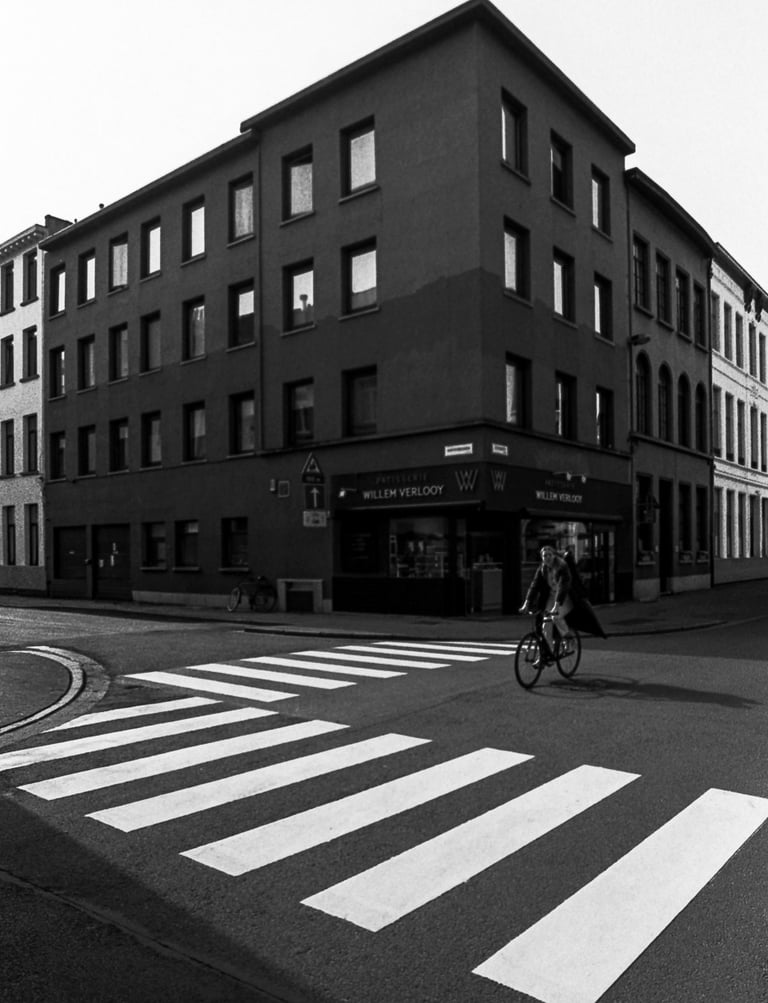 Black and white street photography of a cyclist crossing a zebra walk near a corner building in Antwerp.