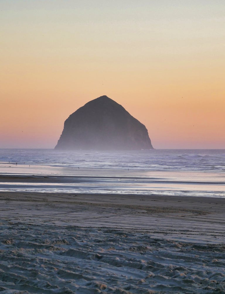 A wide sandy beach at sunset featuring the iconic Haystack Rock monolith off the Oregon coast.