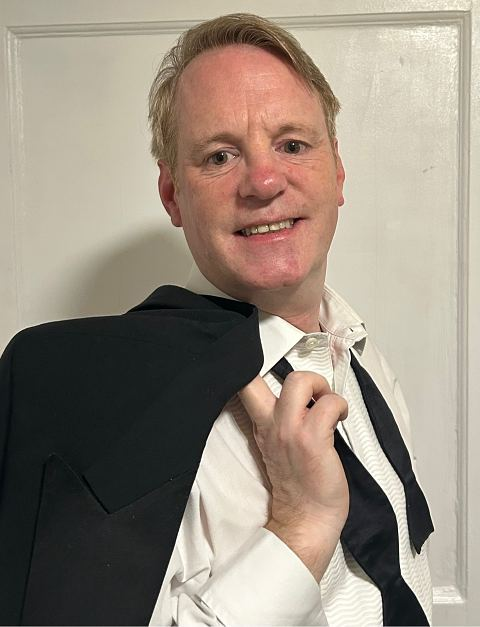 A smiling man in a white dress shirt with an untied black bowtie holding a tuxedo jacket over his shoulder.