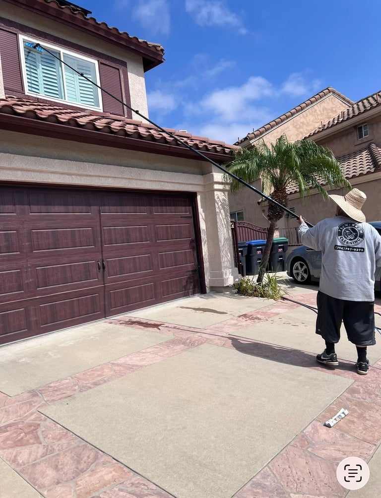Professional window cleaner using a water-fed pole to wash second-story exterior windows on a residential home.