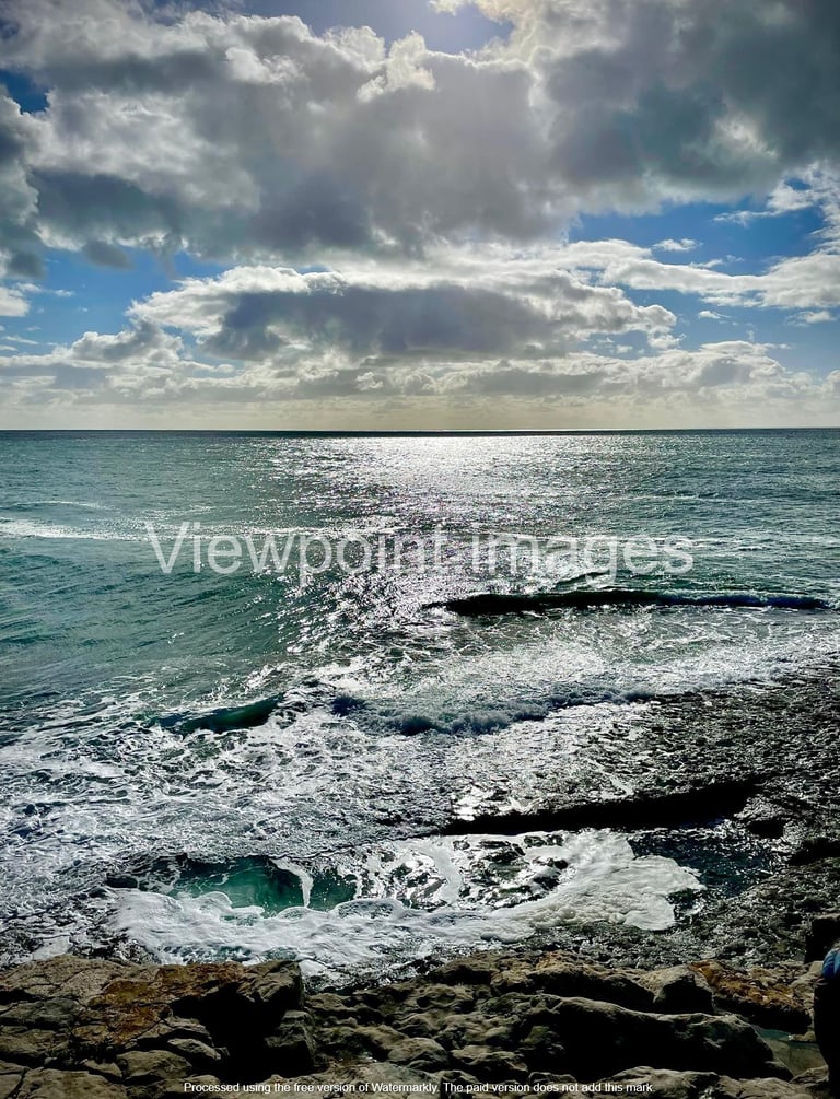 Sparkling blue ocean waves crashing against a rocky shore under a cloudy sky with bright sunlight.