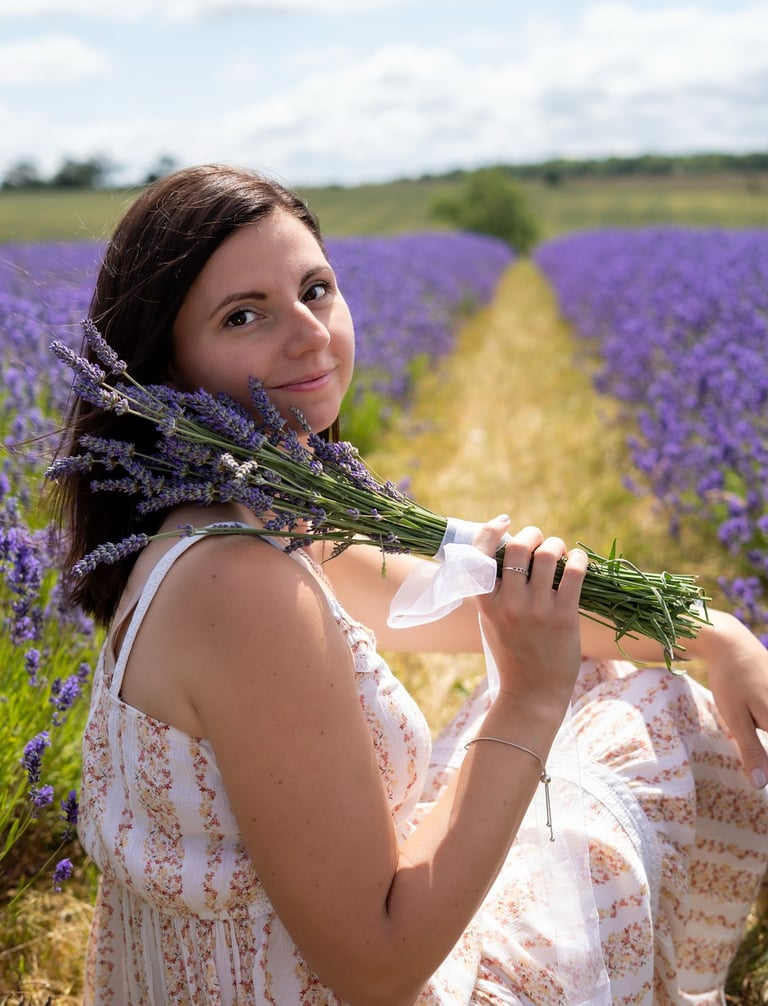 Woman sitting in a lavender field, wearing a cream flowing dress and holding a bunch of lavender