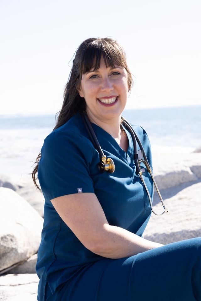 Smiling female healthcare professional wearing blue scrubs and a stethoscope at the beach.