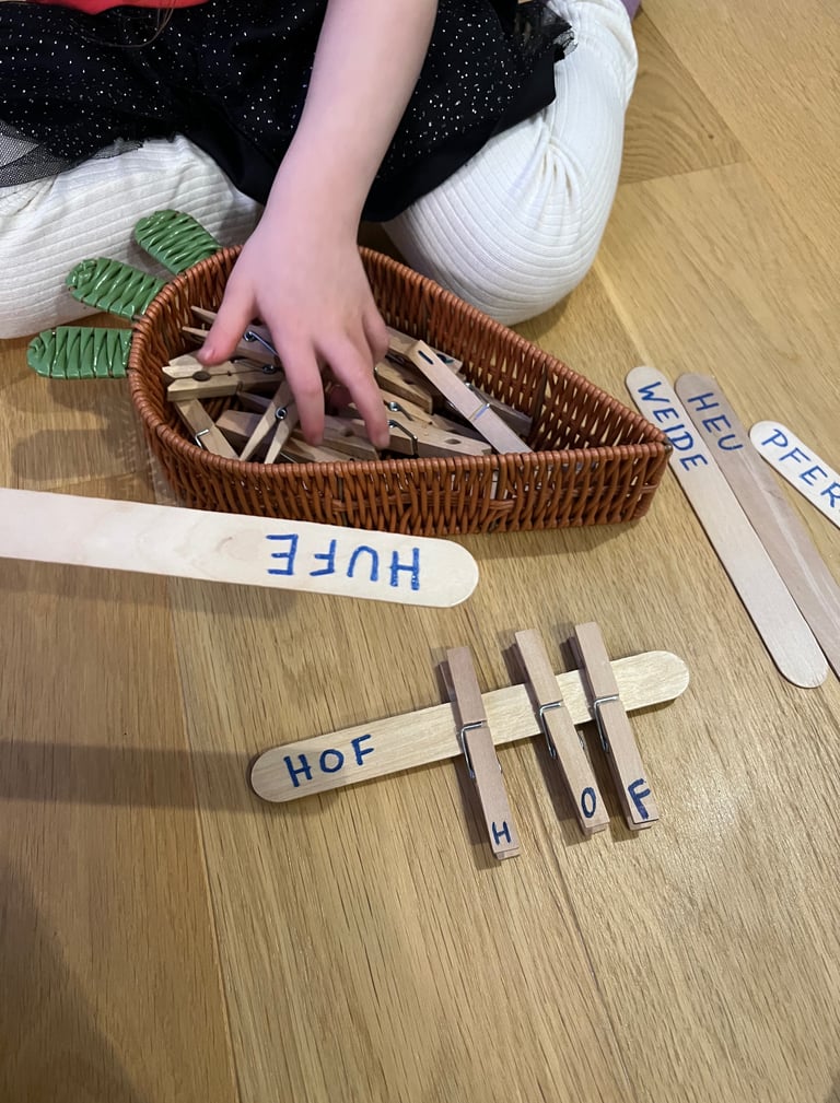 Child using wooden clothespins to match letters on craft sticks for a German word spelling activity.