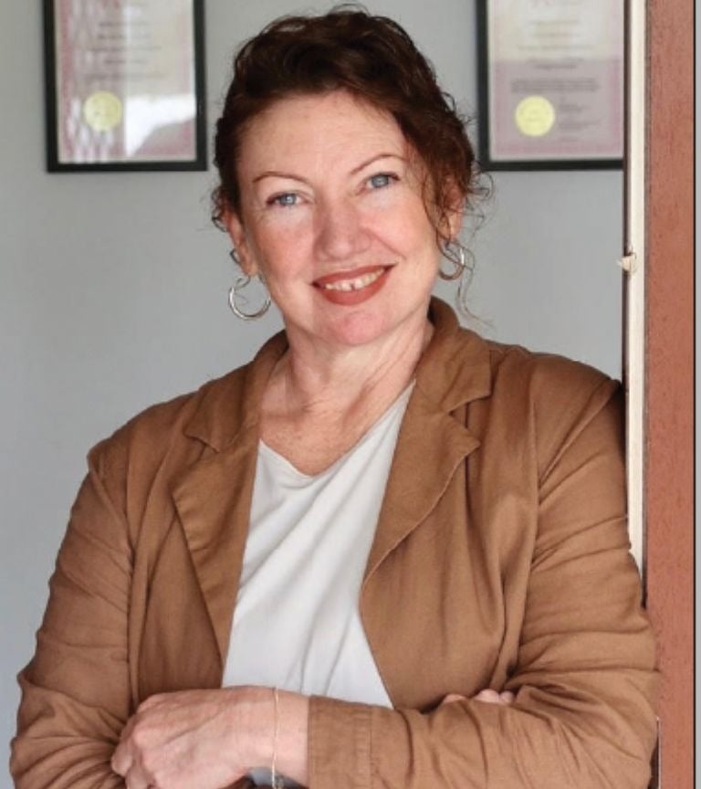 A smiling professional woman with curly dark hair wearing a brown blazer leaning against a doorway.