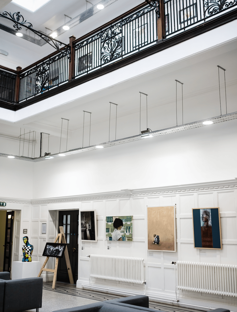 Paintings in a gallery with sofas on ground level underneath a balcony.  Partial glass ceiling. 