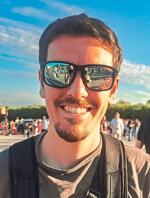 Portrait d’un homme souriant avec des lunettes de soleil reflétant une foule, ciel bleu et ambiance ensoleillée d’événement