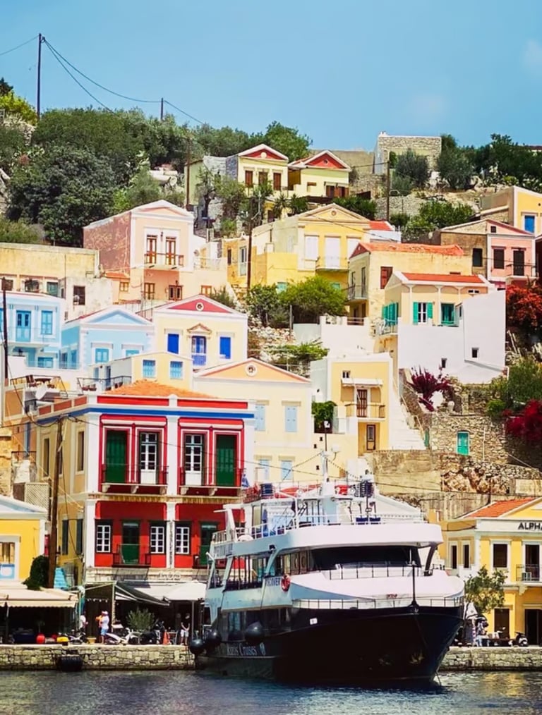 Colorful neoclassical houses on a hillside overlooking a cruise boat in Symi Island harbor, Greece.