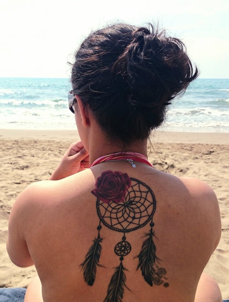 Mulher de costas na praia, com tatuagem de rosa e filtro dos sonhos, símbolo de memória ancestral