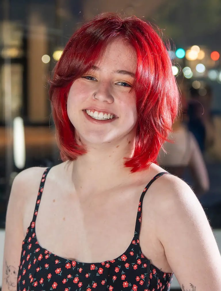 Smiling young woman with vibrant red bob haircut wearing a floral sundress in a city setting.
