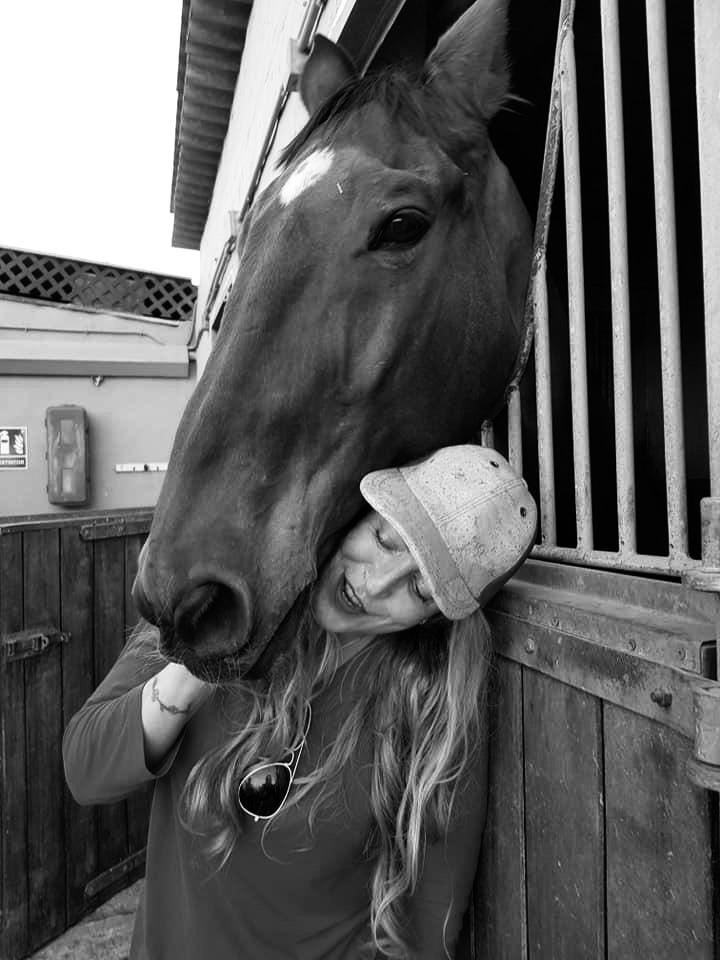 A smiling woman wearing a baseball cap leans against a brown horse standing in a stable stall.