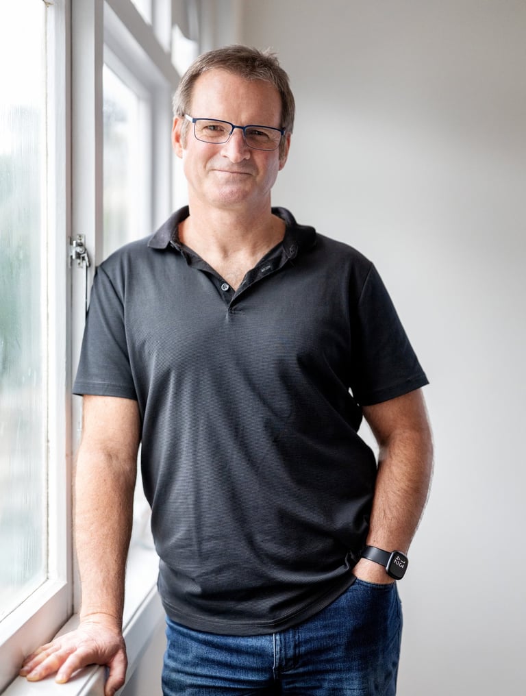 Photo of a man wearing a glasses, a black polo shirt and jeans, leading on a window sill and smiling