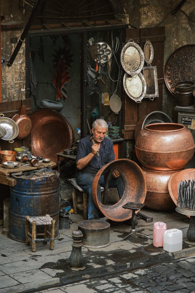 A skilled coppersmith hammers a large copper bowl in a traditional artisan workshop filled with metalware.