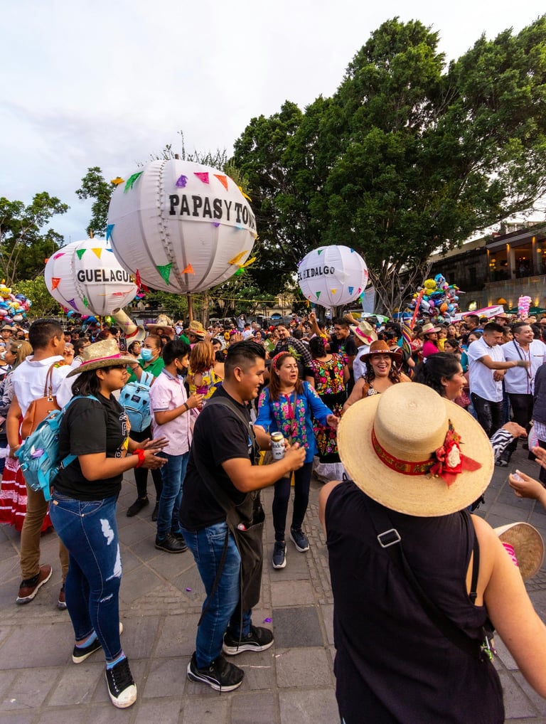 Gente bailando en calenda de Guelaguetza en Oaxaca con globos gigantes y música de fondo
