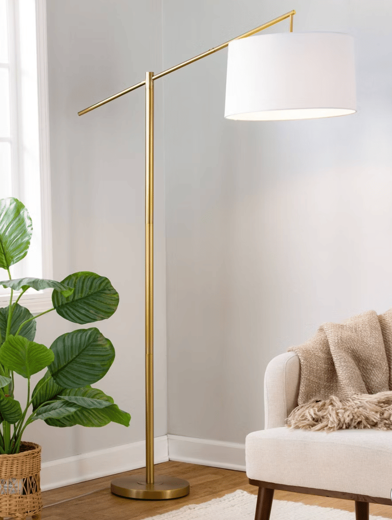 A modern gold arched floor lamp with a white drum shade standing next to a cream armchair.