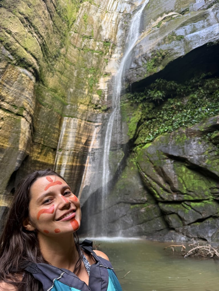 Une femme souriante, le visage peint selon la tradition indigène, se tient devant une haute cascade 