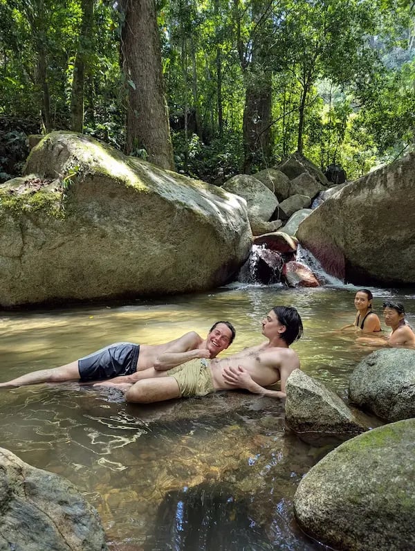 Friends relaxing in a natural jungle swimming hole with a small waterfall and large granite boulders.