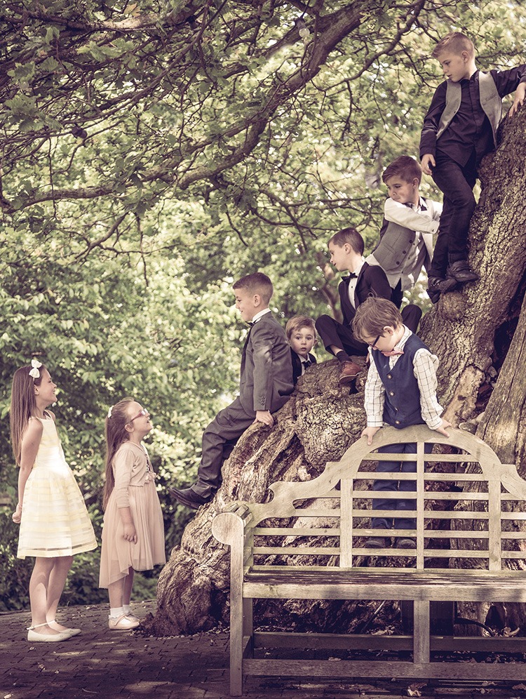 Young wedding guests in formal attire and flower girl dresses posing on a large tree outdoors.