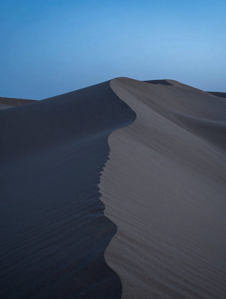 Clean, modern photography of desert sand dunes at twilight, focusing on the elegant curves and sharp shadows, deep blue and slate grey tones, minimalistic and serene aesthetic.
