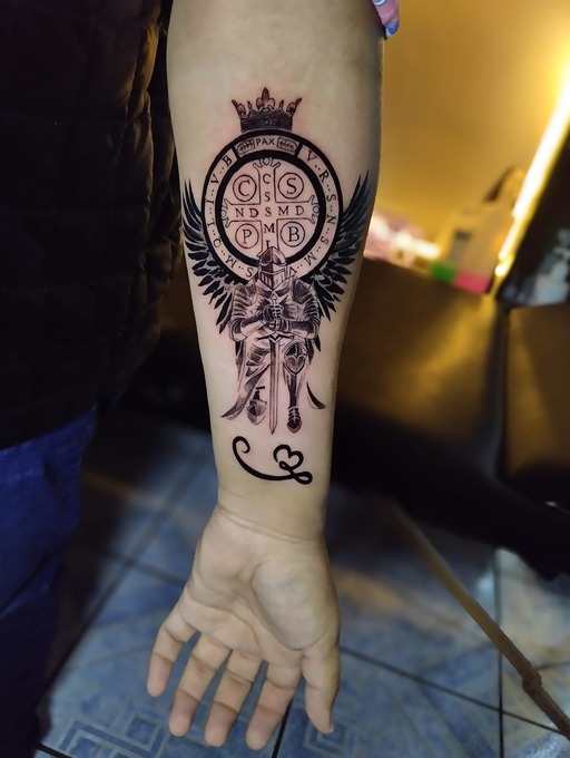 Tatuaje detallado en tinta negra en el antebrazo que presenta una medalla de San Benito: