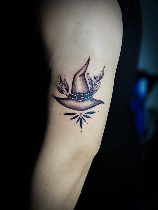 Tatuaje de sombrero de bruja en tinta negra en el brazo de una persona, con hojas botánicas.
