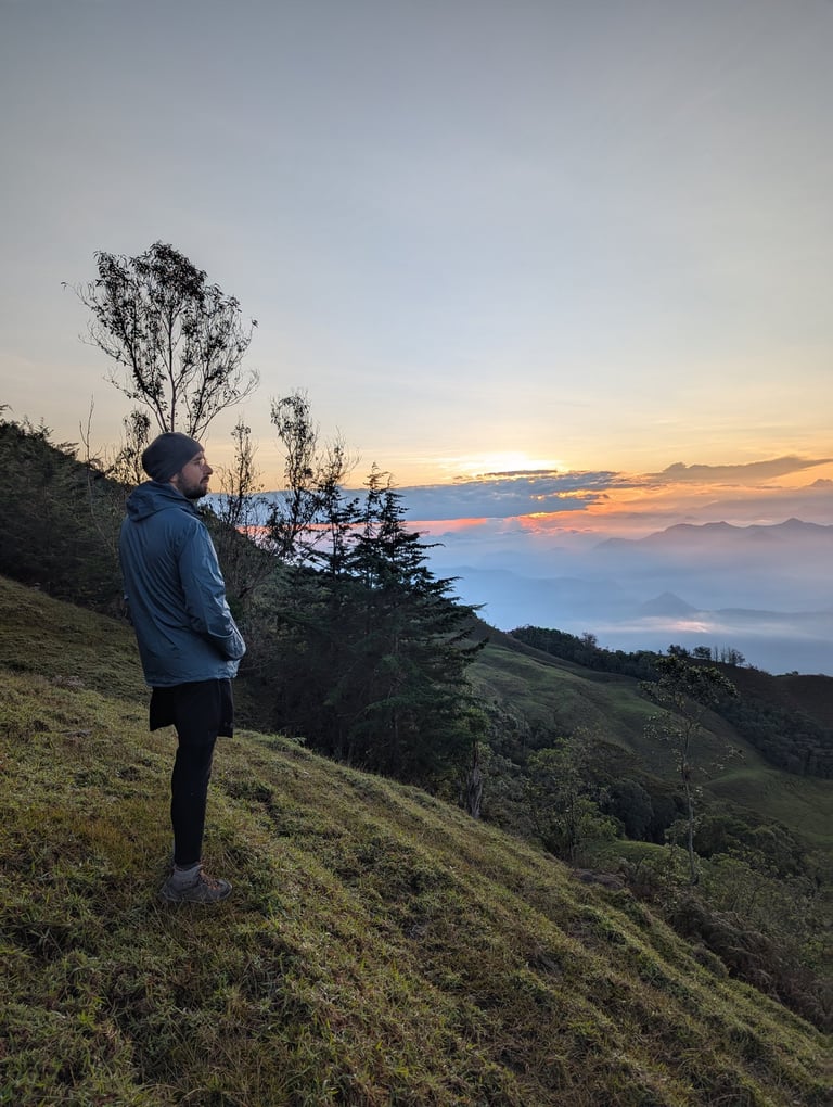 A male hiker standing on a grassy mountain ridge watching a scenic sunrise over a misty valley.