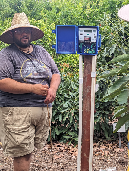 Mira Costa student explaining his water sensor project, standing next to a blue box on a wooden post